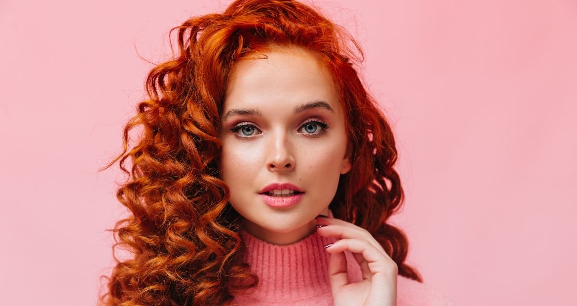 Menina com blusa rosa de gola alta com cabelo cacheado em tons de ruivo em um fundo rosa.